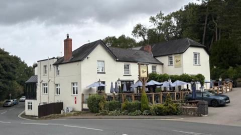 The exterior of a hotel and outdoor eating area. The hotel has a cream facade with a black tiled roof. It has seating on a raised level under beige umbrellas with glass panels in between wooden posts forming a fence. In front of the raised seating area is a lower seating area. There is a large pub sign in among plants and bushes in a garden border. To the right of the building is the entrance to the hotel carpark. A road runs alongside the hotel. 