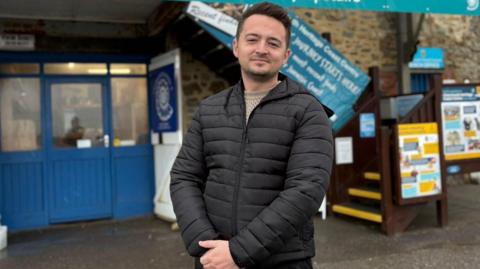 Ryan Eddowes stands outside Charmouth Heritage Coast Centre (Dorset) where his grandparents would take him as a youngster, fostering a love of animals. He has short black hair, black stubble and wears a black, puffy jacket with his hands clasped in front of him.