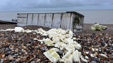 A shipping container washed up on the shingle beach, with foam at the forefront of the photo. It is a grey day.