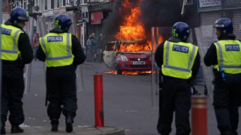 Four police officers in riot helmets stand in a line with their backs to the camera, beyond them a red car is on fire