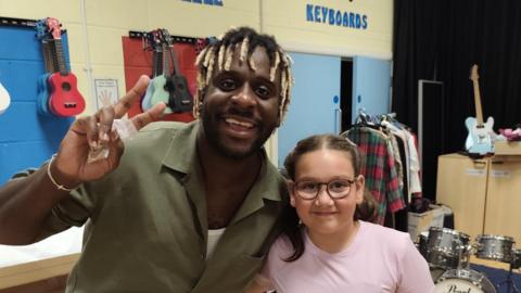 Myles Smith poses and does a peace sign with his hands as he stands next to Matilda. Smith wears a green shirt with white vest underneath and Matilda wears a pink top and black shorts.