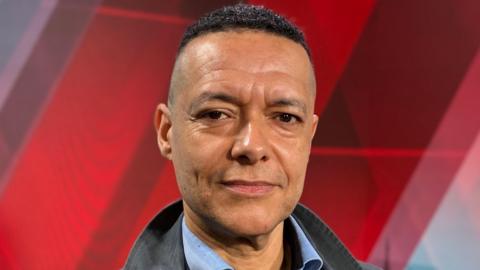 Clive Lewis stares at the camera. He is wearing an open necked blue shirt and a black jacket.