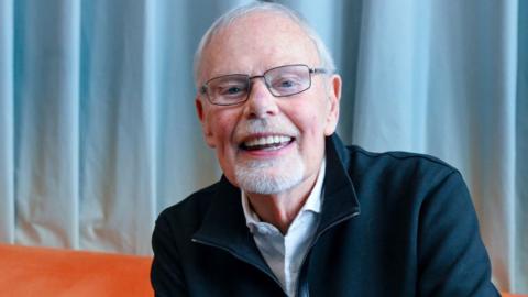 Bob Harris sitting on an orange sofa against a light blue curtain, in March 2026