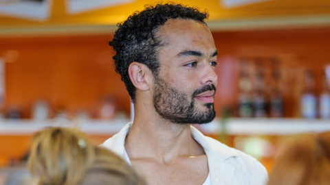 Matthew Punter, looking to the right. He is wearing a white top and yellow shirt, and two blurred ladies are in the foreground.