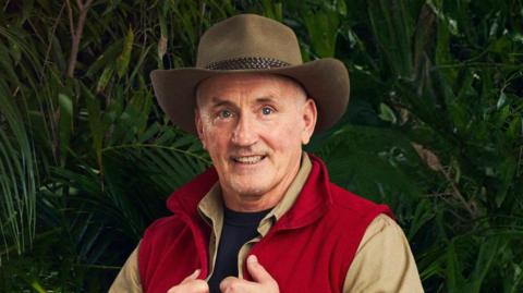 Barry McGuigan standing with a red body warmer and red shorts and a brown hat on, he is pictured with his hands holding on to the front of his body warmer. he is standing beside a jungle. 