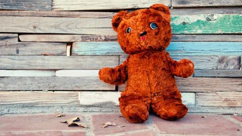 A brown teddy bear propped up against a wooden wall