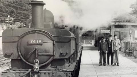 A black and white image of a steam train waiting on a platform while three men have their arms round each other smiling for a photo. They are wearing suits and hats, that look like they work at the station.