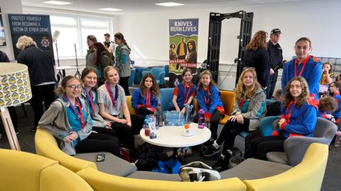 Eight Guides look at the camera while sitting on chairs in a semi-circle shape with posters, chairs, a lampshade and other Guides and police officers in the background.