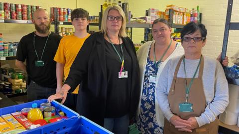 Volunteers at Gleadless Valley Food Bank stood in a storage room filled with shelves stocked with non-perishable food, such as canned goods.
