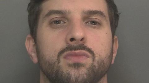 A mugshot of Thomas Cashman, who has dark brown hair and black stubble, and purses his lips at the camera with a defiant expression