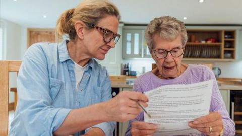 A woman and her daughter sit at a table in the kitchen and look through their bills looking concerned. 