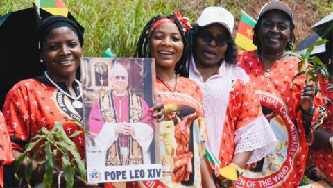 A crowd of women dressed in specially designed red fabric for the Pope's visit in Bamenda. One holds a poster of Pope Leo several others hold branches of green leaves.