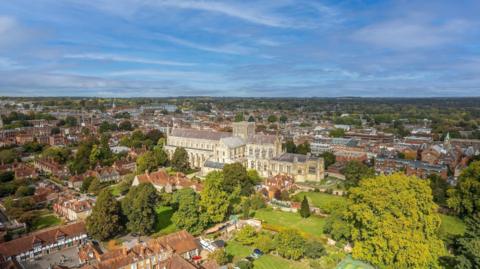 An aerial view of Winchester. The large stone cathedral building is surrounded by homes. In the foreground there is a large tree park area with trees.