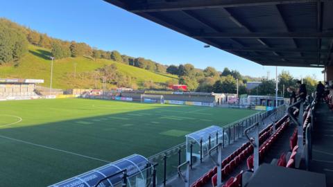 A football stadium with red seating overlooking an artificial‑turf pitch, with green hills in the background.