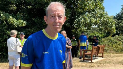 An old man wearing a blue football shirt with a yellow neckline and sleeve rings smiles slightly as he stands in a scorched field. There are other people stood in the background and there is a new, brown wooden bench. There are hedges and trees bordering the field.