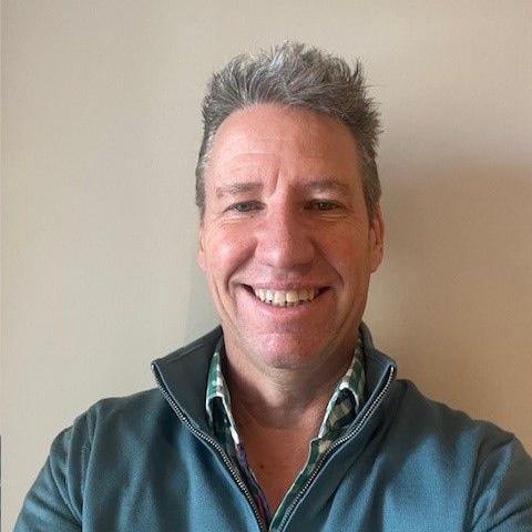 Andy Barton smiles at the camera - he is wearing a blue zipper top over a green and white check shirt and stands against a cream-coloured wall