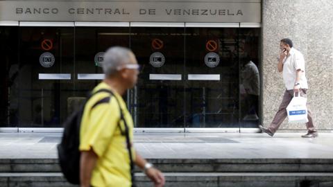A person wearing a yellow T shirt and a backpack walks past the central bank of Venezuela as another person in a white shirt walks holding a shopping bag in the background, outside a glass entrance with the words 'Banco Central de Venezuela' on it, in Caracas in 2017.