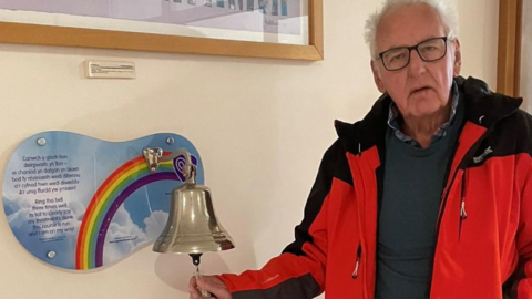Noel, wearing a red and black rain coat, ringing a metal bell in a hospital corridor. There is a rainbow mural on the wall the bell is attached to. 