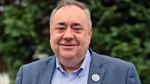 Alex Salmond smiles at the camera. He has grey hair and wears a blue and white shirt and a grey blazer with an Alba pin. He appears to be standing in front of greenery.