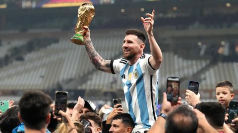 Lionel Messi wearing an Argentina shirt holding aloft the World Cup trophy in a football stadium
