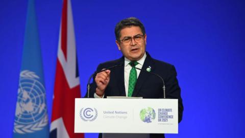 Honduras President Juan Orlando Hernández speaking at a podium, wearing a black suit and a green dotted tie. Behind him is a blue backdrop with a UN flag and a UK flag to his left. He has short hair and wears square-framed eye glasses. 