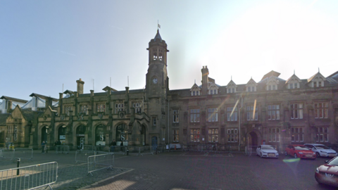 Carlisle railway station which is a grand, three-storey stone building. A central turret has a clock on it. The doorways are arched. Cobbled paving stones are seen at the front.