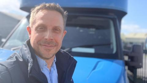 A man with short, grey hair smiles and takes a selfie. He has a blue shirt and a dark coat on. He stands in front of a blue van.