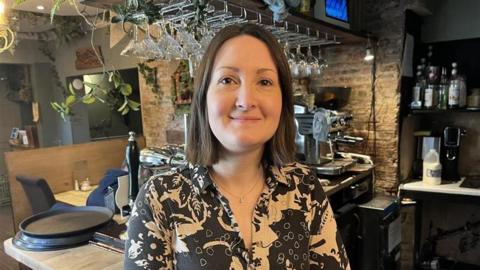 A woman in a patterned shirt stands smiling behind a cosy bar counter, with hanging glasses, bottles and a coffee machine in the background.