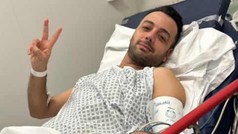 Pouria Zeraati lies in a hospital bed, wearing a hospital gown and wrist band, looking at the camera with two fingers raised in a peace sign