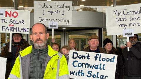 Protesters holding up placards opposed to solar farm development in front of glass doored entrance to Cornwall Council