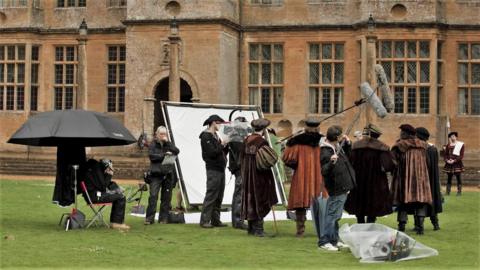 A group of actors dressed in Tudor clothing standing the the grass outside Montacue House, which is a large Elizabethan mansion made from stone. There is a film crew holding microphones above the actors and a large light reflector sheet is positioned behind the camera.