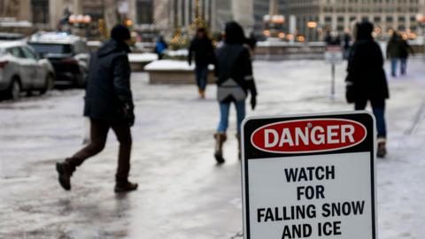 People walking on icy road and pavement near a danger sign that warns of falling snow and ice.