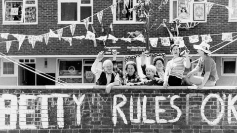 A group of Fulham residents show their support for Queen Elizabeth II's twenty-fifth year of reign by waving enthusiastically under strings of banners.