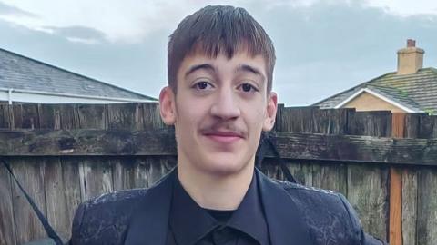 Taylor Charlton is a young man standing outdoors in front of a wooden fence. He is wearing a dark buttoned shirt and a dark patterned jacket. He has short dark hair and a light moustache. Behind the fence are the roofs of nearby houses and a cloudy sky.