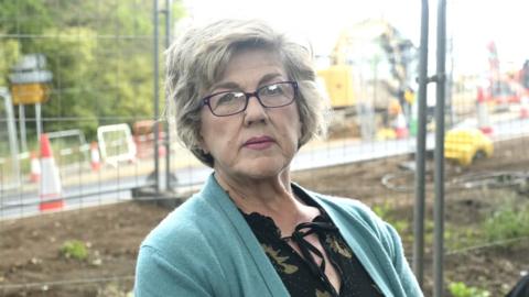 A woman with grey hair and glasses looks at the camera. She is wearing a green cardigan and black top. The background of the image is a muddy area sealed off by wire metal fencing, with traffic cones dotted around and a digger in the distance.