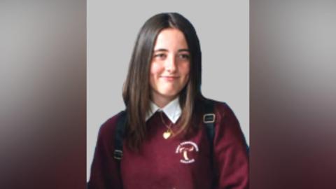 An image of a girl, wearing a burgundy school uniform and white shirt. She has long brown hair. There is a grey wall behind her. 