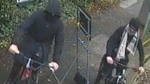 The grainy image shows a person with a black face covering and a thick black puffer coat on a red and black bike next to a person dressed all in black with a white patterned scarf on a black bike.