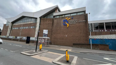 Outside view of Coventry Sports and Leisure Centre on Fairfax Street. A road runs in front of a brick building with large glass windows at the top and a jagged roof. The sign identifying the building as 'Coventry Sports and Leisure Centre' is on the brick wall.

