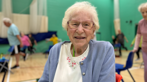 Lillian has short white hair and is smiling and wearing glasses, in a white blouse and lilac cardigan. She is sitting in a sports hall with other people exercising in the background.