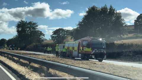 Firefighters and a fire engine are at the side of the road and charred vegetation can be seen.