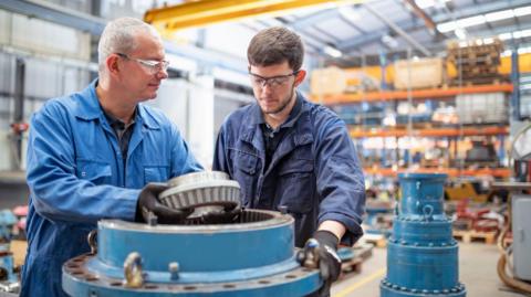 Two men, wearing protective goggles and blue overalls, inspecting a blue, metal pump in a factory.