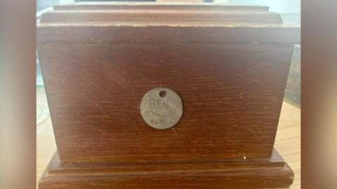 A brown casket of ashes box with the name Ben written on it, along with the numbers 848679.