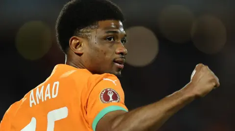 Amad Diallo, seen from behind wearing an orange Ivory Coast shirt, clenches his right fist in celebration