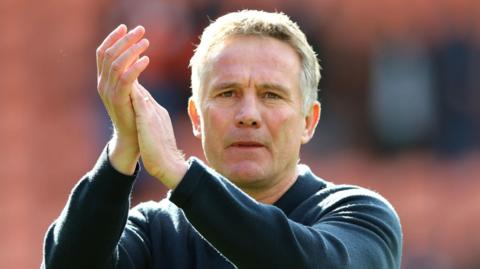 Phil Parkinson clapping after a match in front of a blurred background. He is wearing a navy blue polo shirt and looking towards the camera.
