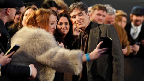 Cillian Murphy, in a grey jacket, is standing on a red carpet queue smiling for a selfie, which is being taken by a woman in a white furry coat