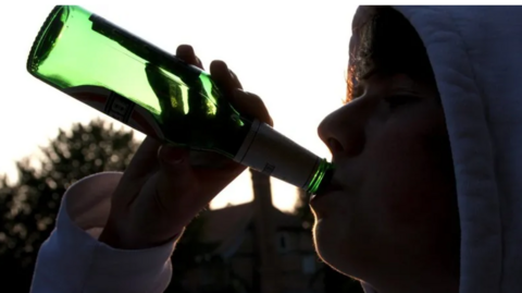 An anonymous young person in a grey hoodie drinking beet straight out of green bottle