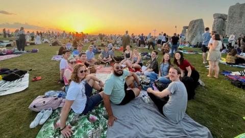 A group of young people sitting on a blanket on some grass next to Stone Henge. The sun is setting and there are lots of other people around them.