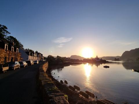 Image of Plockton, Highland showing a road to the left of the image with houses along it. The sun is very low in the sky shining on the water in front of it.