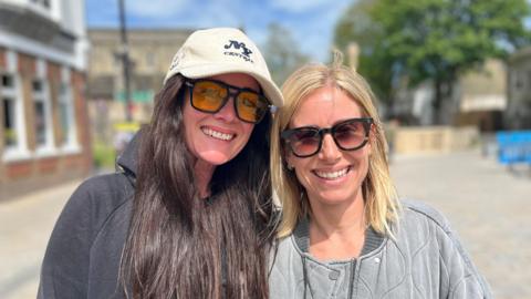 Emilie on the left has long dark brown hair and wears a cap and sunglasses. On the right Melanie has shoulder-length blond hair and wears sunglasses. 