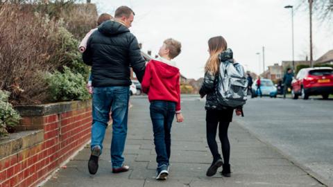 Father with 3 kids walking along the pavement, walking away from the camera. They are on a residential street in the UK 